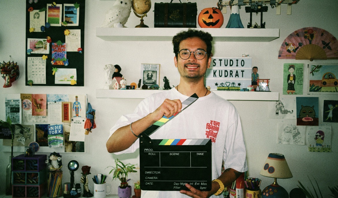Kudrat holding a clapperboard in his studio, surrounded by art and objects, with a letterboard reading Studio Kudrat